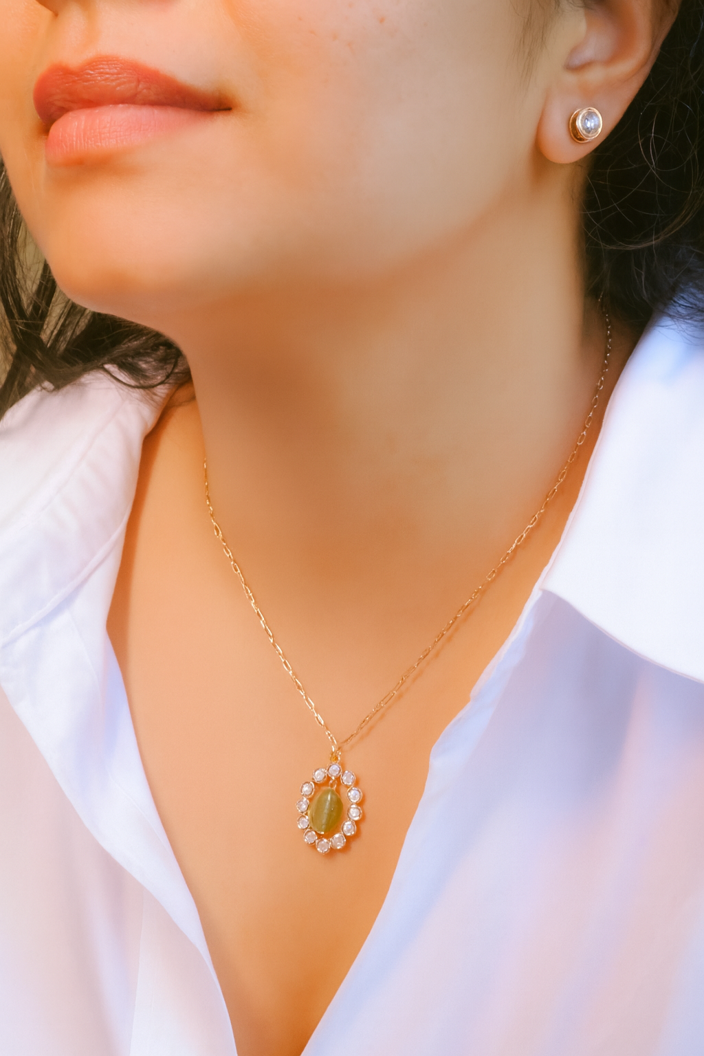 Close-up of a person wearing a gold necklace with a green pendant and white stones, against a blurred background.