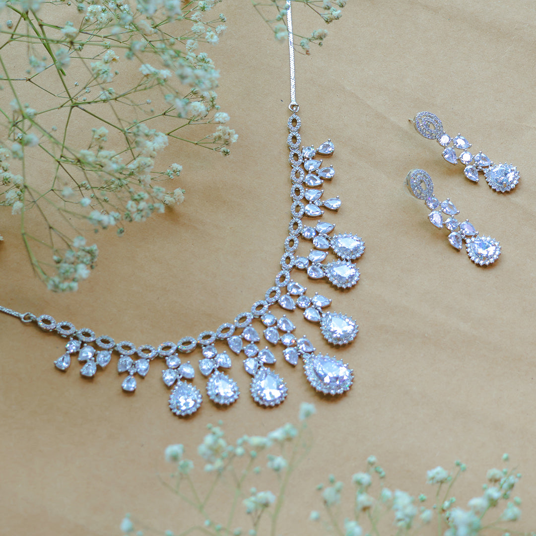 Silver necklace and earrings with clear gemstones on a beige background with small flowers.
