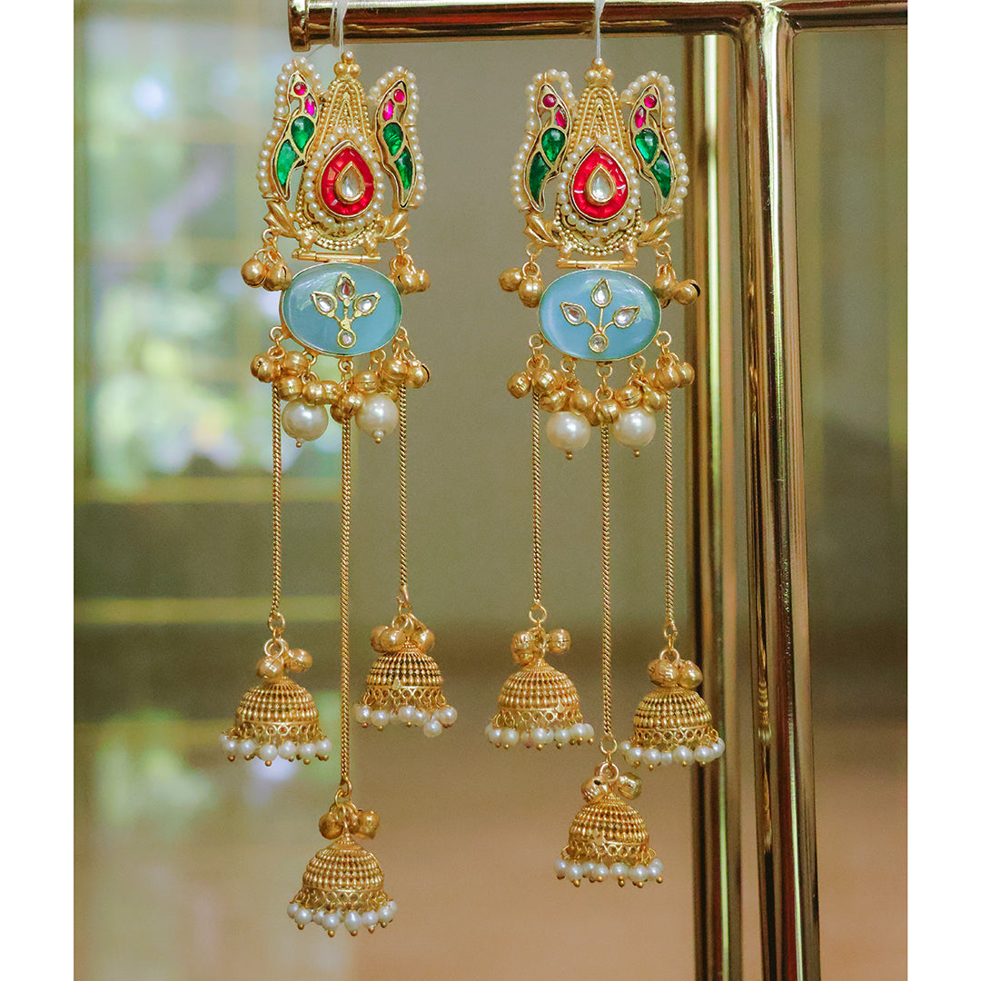 Ornate gold earrings with colorful stones and pearl-like beads on a reflective surface.