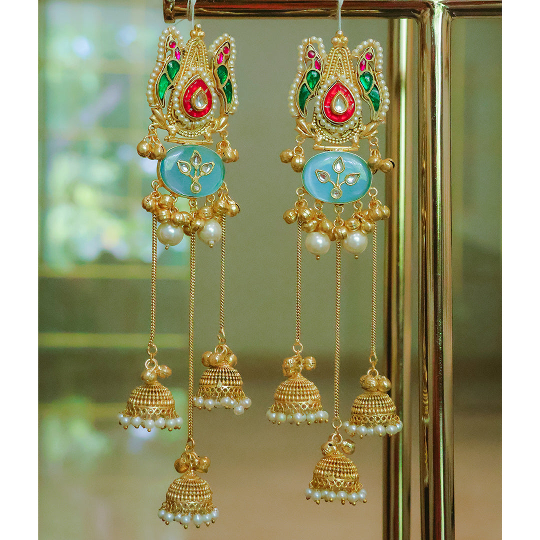 Ornate gold earrings with colorful stones and pearl-like beads on a reflective surface.