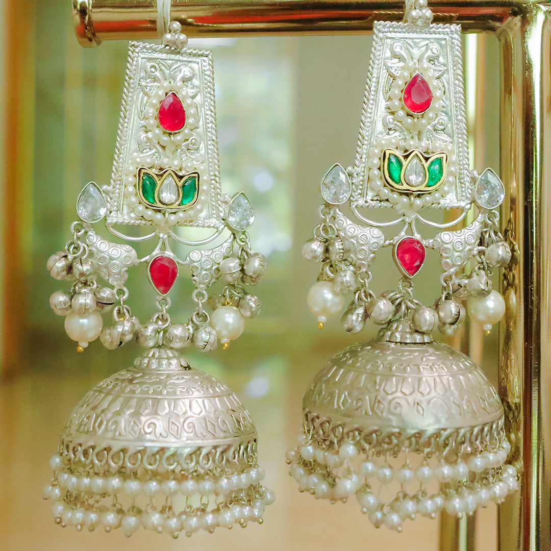 Ornate silver earrings with red and green stones and pearl-like beads on a reflective surface.