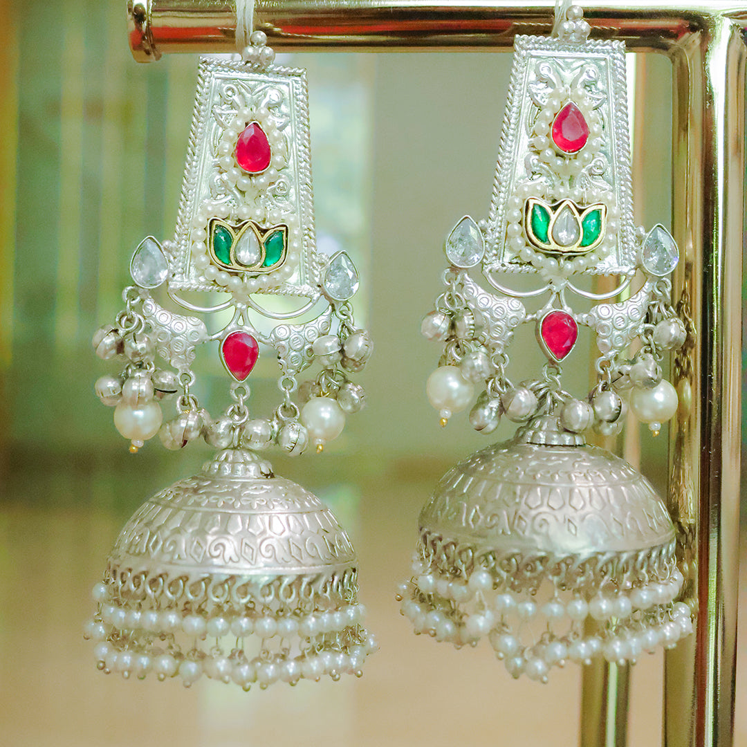 Pair of ornate silver earrings with pearls and gemstones on a reflective surface.