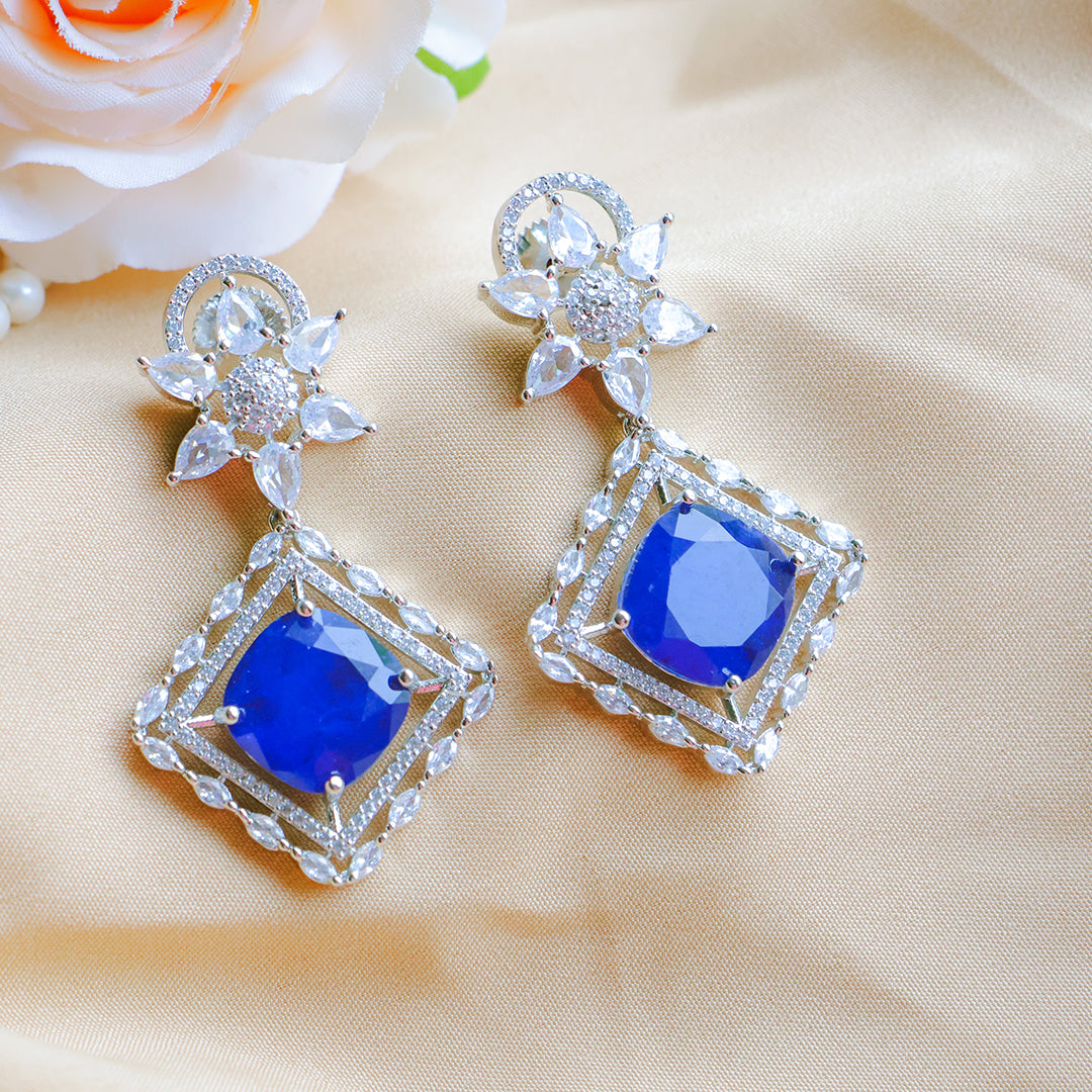 Pair of blue gemstone earrings with silver accents on a beige fabric background.