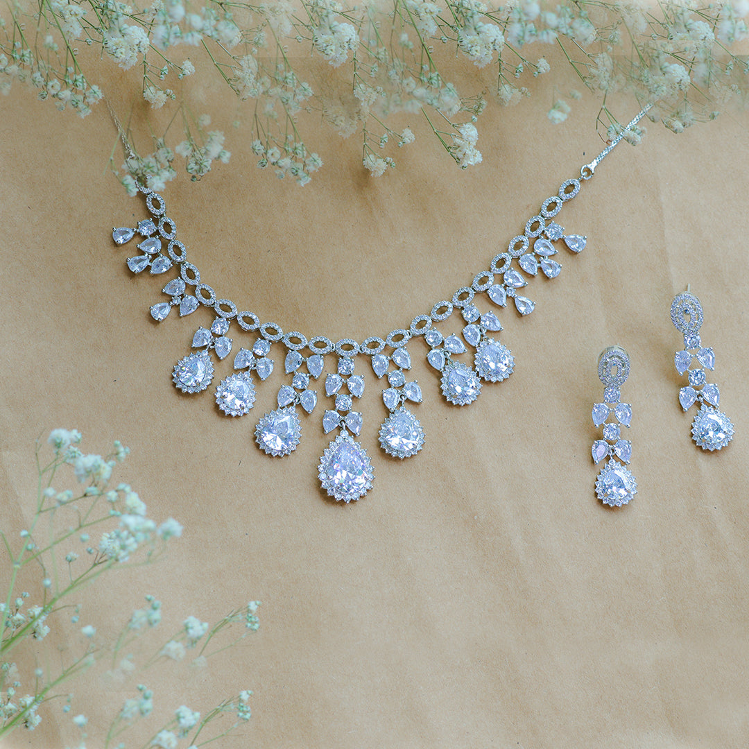 Silver necklace and earrings with clear stones on a beige background with small white flowers.