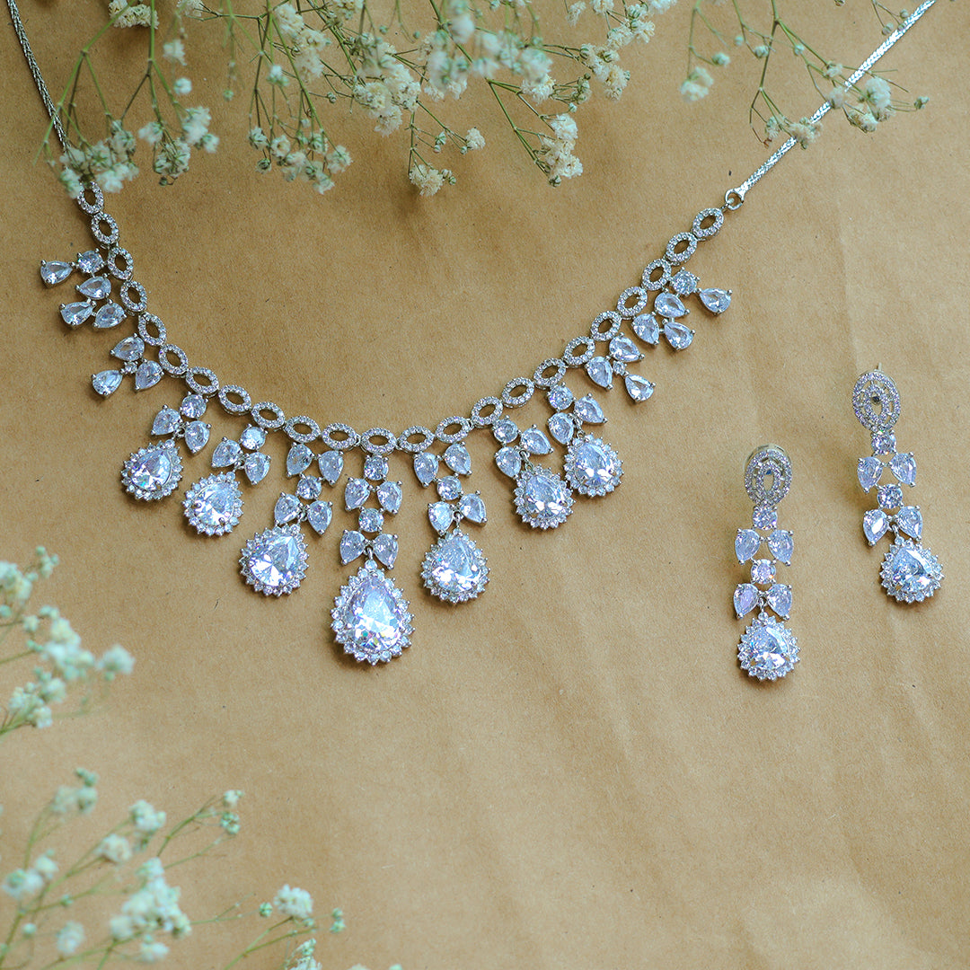 Silver necklace and earrings with teardrop-shaped crystals on a beige background with small white flowers.